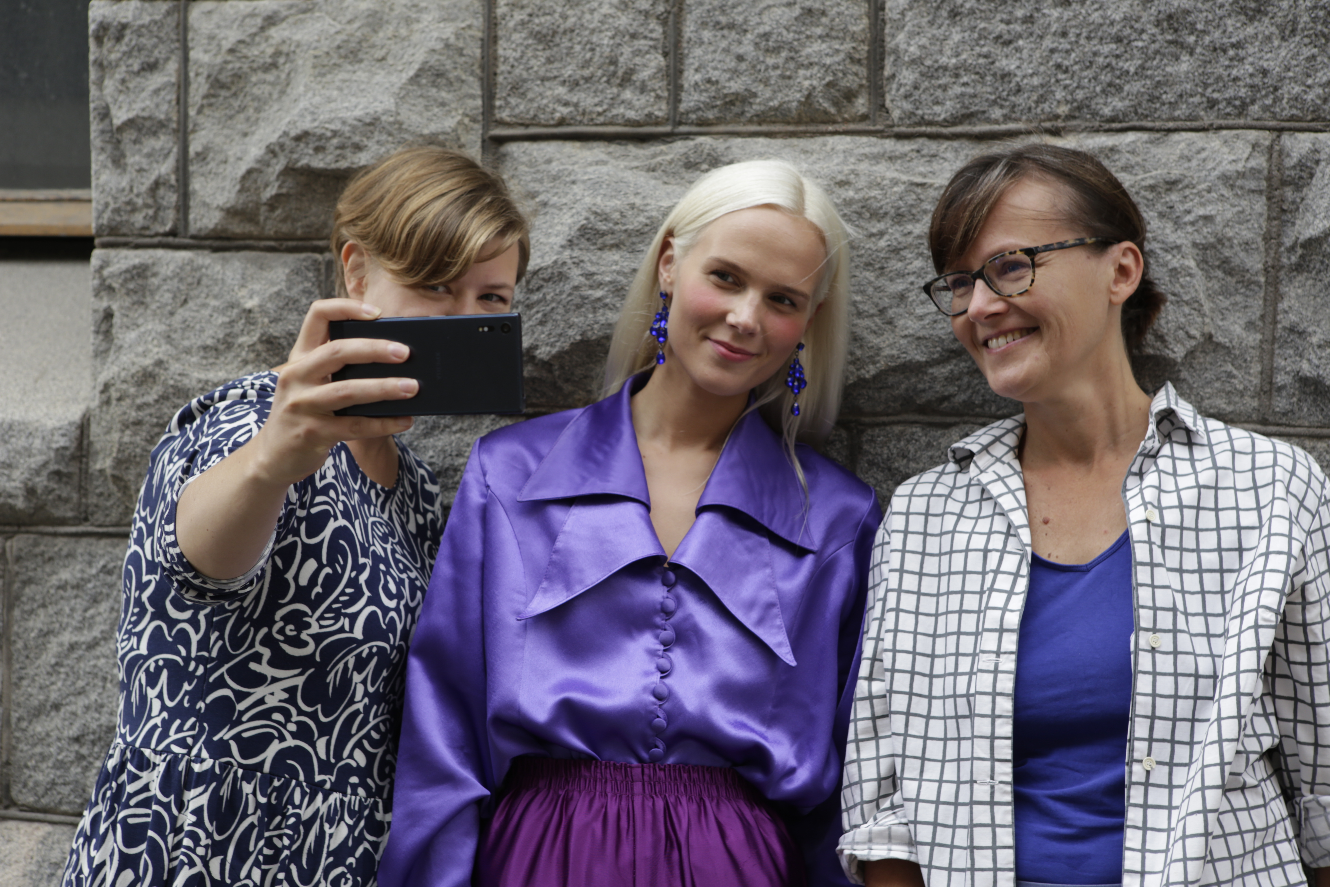 Kolme naista seisoo vierekkäin kiviseinän edessä. Yksi ottaa puhelimella selfietä kaikista kolmesta.