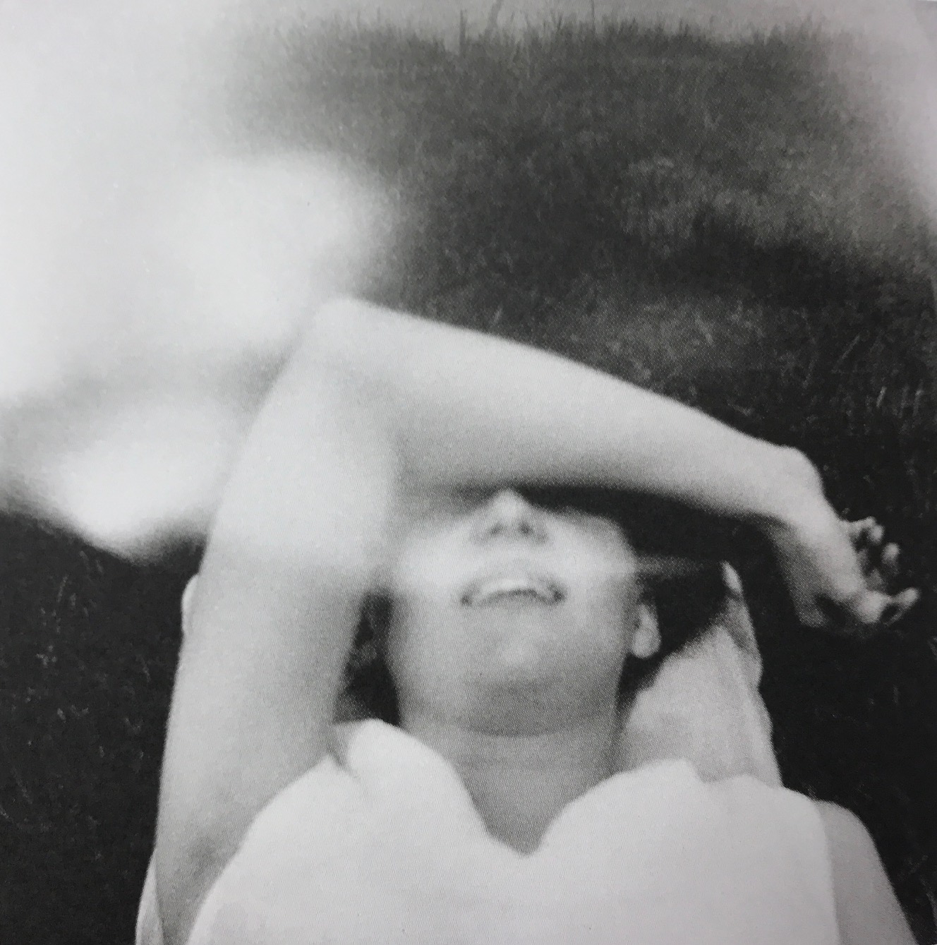 Mustavalkoisessa kuvassa nainen makaa nurmikolla selällään käsivarsi silmien päällä. Kuvan yläreunassa on vaaleita sumuisia kohtia. 