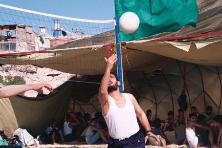 Mies lyö lentopalloa verkon yli. Hänen takanaan teltassa istuu useita ihmisiä.