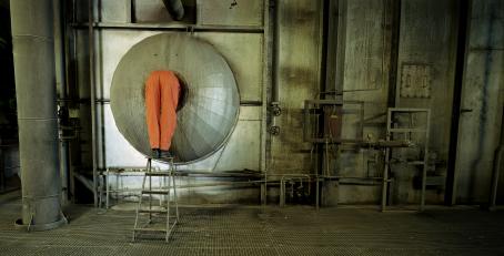 Näkymä tehtaasta. Vasemmassa reunassa oranssihaalarinen henkilö seisoo jakkaran päällä kurkistamassa putkeen, ja hänestä näkyvät vain jalat.