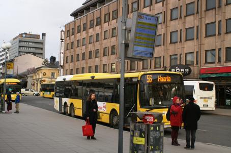Keltainen Turun paikallisbussi numero 56 pysähtyneenä pysäkille. Sen vieressä seisoo ihmisiä. Taaempana näkyy lisää busseja ja rakennuksia.