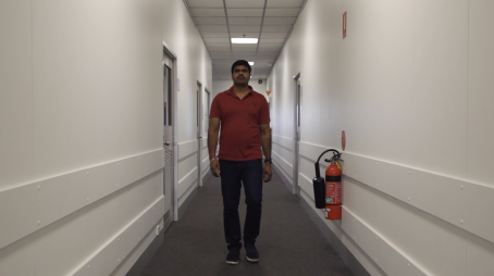 Punapaitainen, tummahkoihoinen mies kävelee käytävällä kameraa kohti. Miehen kulmat ovat kurtussa. Käytävällä on ovia molemmilla seinillä ja seinällä on myös vaahtosammutin. 