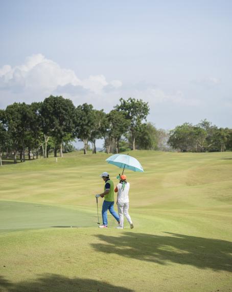 Vihreällä golfkentällä kävelee kaksi henkilöä. Aurinko paistaa, ja toinen pitää sateenvarjoa tai päivänvarjoa heidän yläpuolellaan.