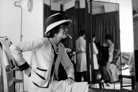Mustavalkoisessa kuvassa Coco Chanel istuu tuolilla ateljeessaan. Hänen suustaan roikkuu tupakka. Takana olevasta peilistä heijastuu ihmisiä, jotka sovittavat vaatetta jonkun päälle.