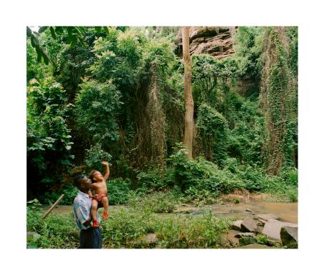 Tummaihoinen mies pitää sylissään tummaihoista lasta, joka huiskuttaa kädellään. Heidän takanaan on vihreää sademetsää ja pieni puro.