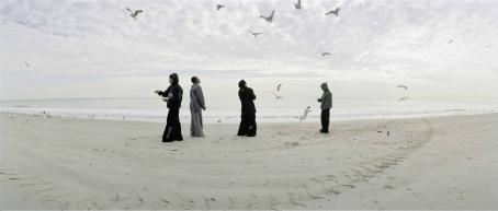 Neljä henkilöä seisoo rannalla ja heidän ympärillään lentää paljon lokkeja. 
