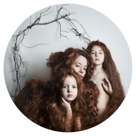 Pyöreässä kuvassa kolme henkilöä, joilla kaikilla on pitkät punaiset hiukset. Hiukset ympäröivät heitä. Takana seinällä on puiden oksia.