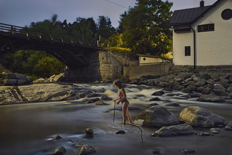 Keskellä jokea seisoo nainen uimapuku päällään. Hänellä on kädessään keppi. Joki on kivinen, ja sen rannalla on talo. Joen yli menee silta.