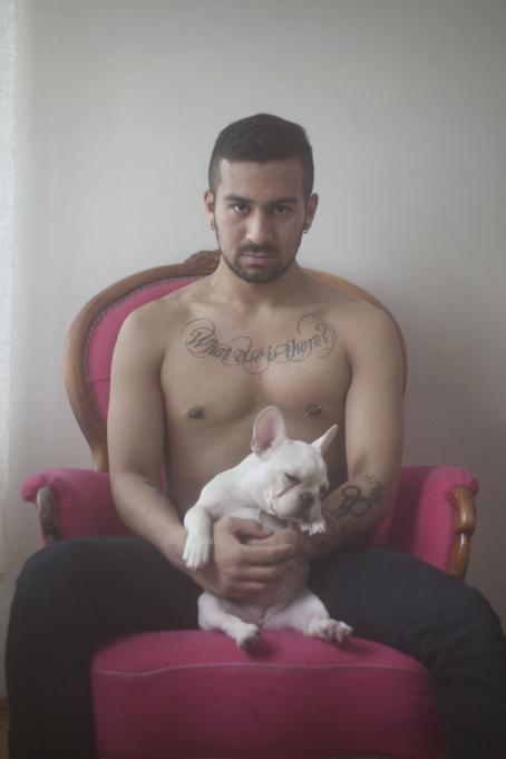 Paidaton mies istuu pinkissä nojatuolissa. Hänen rinnassaan on koristeellinen tatuointi ja hänellä on lävistykset molemmissa nänneissä. Hän pitää sylissään pientä koiraa.