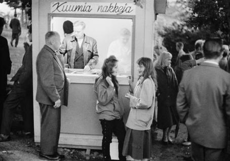 Kioski, jonka seinässä lukee "kuumia nakkeja". Kioskin edessä seisova vanhempi mies on tilaamassa ja hänen vieressään seisoo kaksi nuorta tyttöä. Taustalla näkyy muita ihmisiä. 