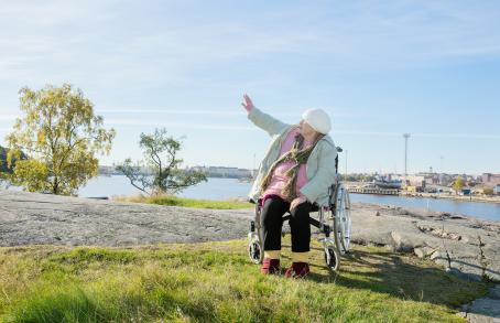Pyörätuolissa oleva vanha nainen rantakalliolla. Taustalla näkyy rakennuksia ja satamaa. Nainen on nostanut kätensä ylös sivulleen ja katsoo samaan suuntaan.