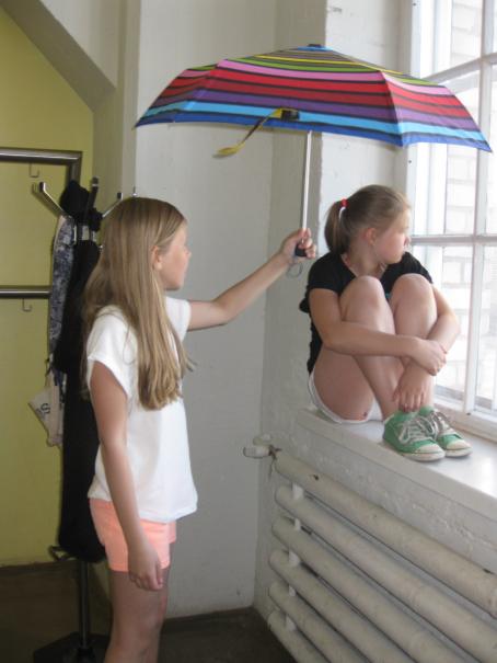 Tyttö istuu ikkunalaudalla polvet koukussa rintaa vasten ja katsoo ulos. Hänen vieressään seisoo toinen tyttö ja pitää värikästä sateenvarjoa ikkunalaudalla istuvan tytön pään päällä. 