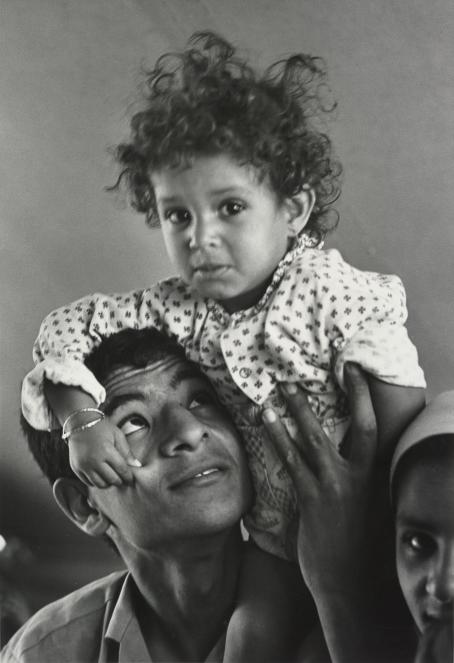 Mustavalkoisessa kuvassa miehen olkapäillä istuu pieni lapsi, joka puristaa miestä poskesta. Mies katsoo ylöspäin lapseen päin.