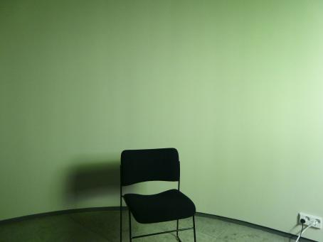 Huoneessa yksi musta tuoli, josta lankeaa varjo seinälle. Seinä on vihreän sävyinen. Seinässä on myös pistorasia, johon on kiinnitettynä kaksi johtoa.