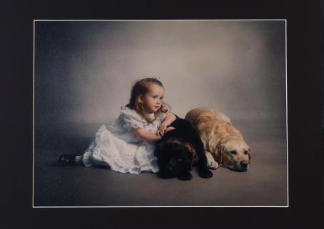 Pieni tyttö vaaleansinisessä mekossa poseeraa lattialla kahden koiran kanssa. 