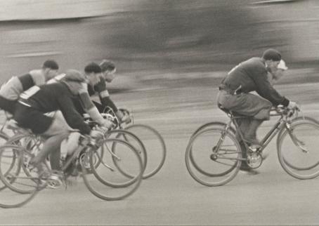 Mustavalkoisessa kuvassa monta henkilöä polkee pyörällä. Tausta on epätarkka. 