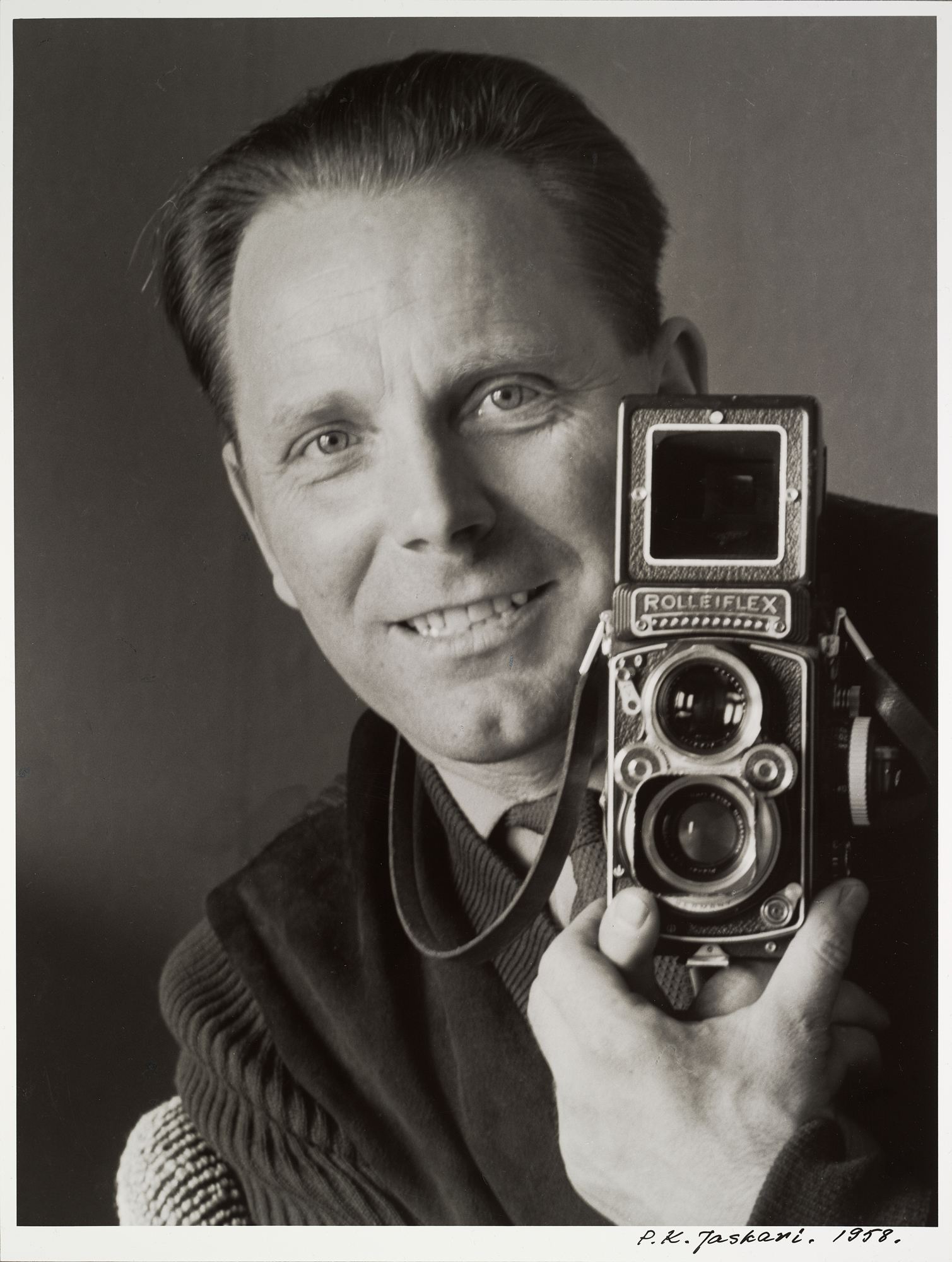 Lehtikuvaaja Helge Heinonen hymyilee kameransa kanssa kollegansa P.K. Jaskarin kuvaamana, 1958. Suomen valokuvataiteen museo. D1999:63/5