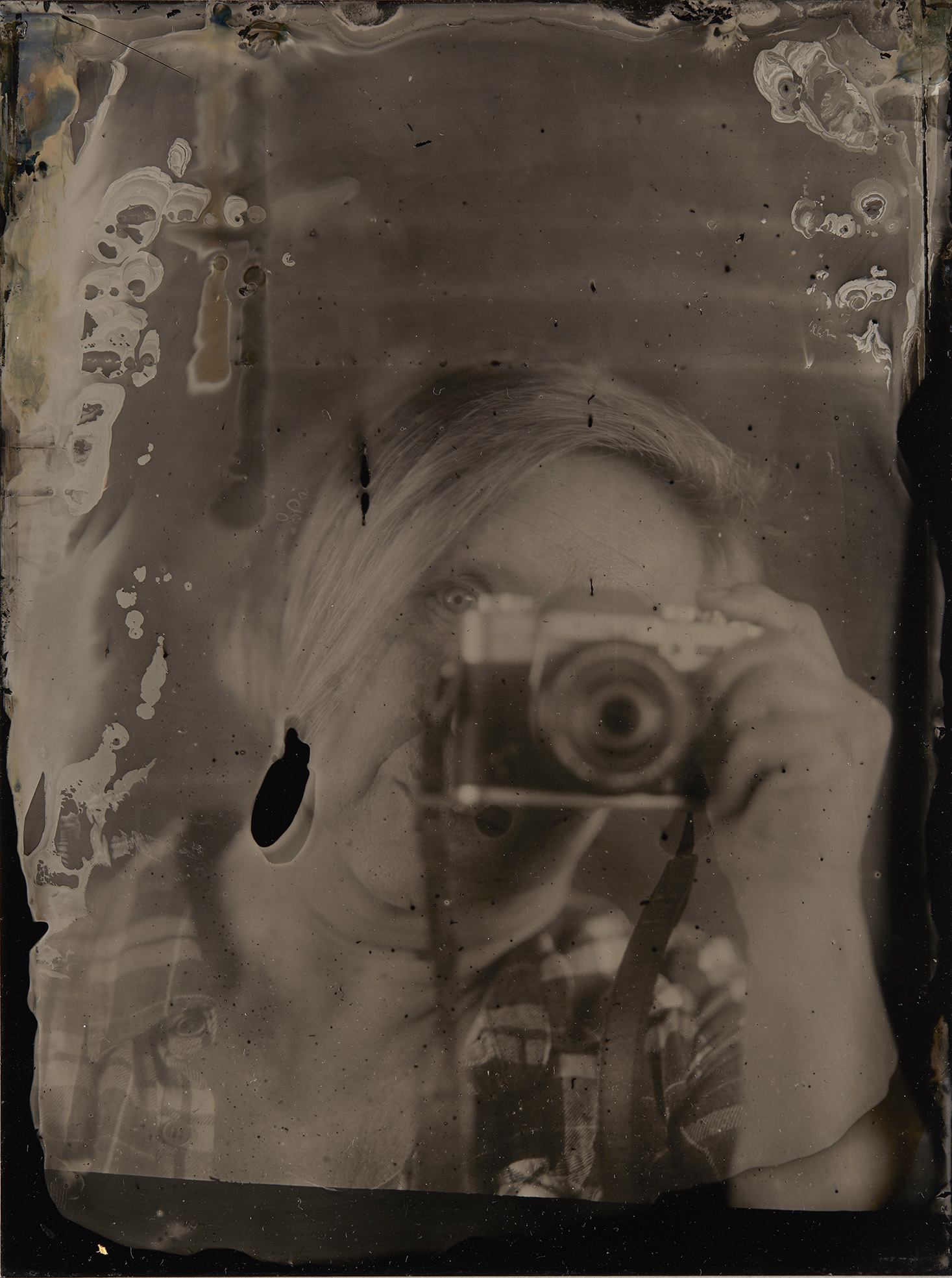 Metallilevylle valotettu mustavalkoinen kuva, jossa näkyvät valoherkkien kemioiden jäljet. Kuva esittää kamera kädessä kuvaavaa henkilöä.