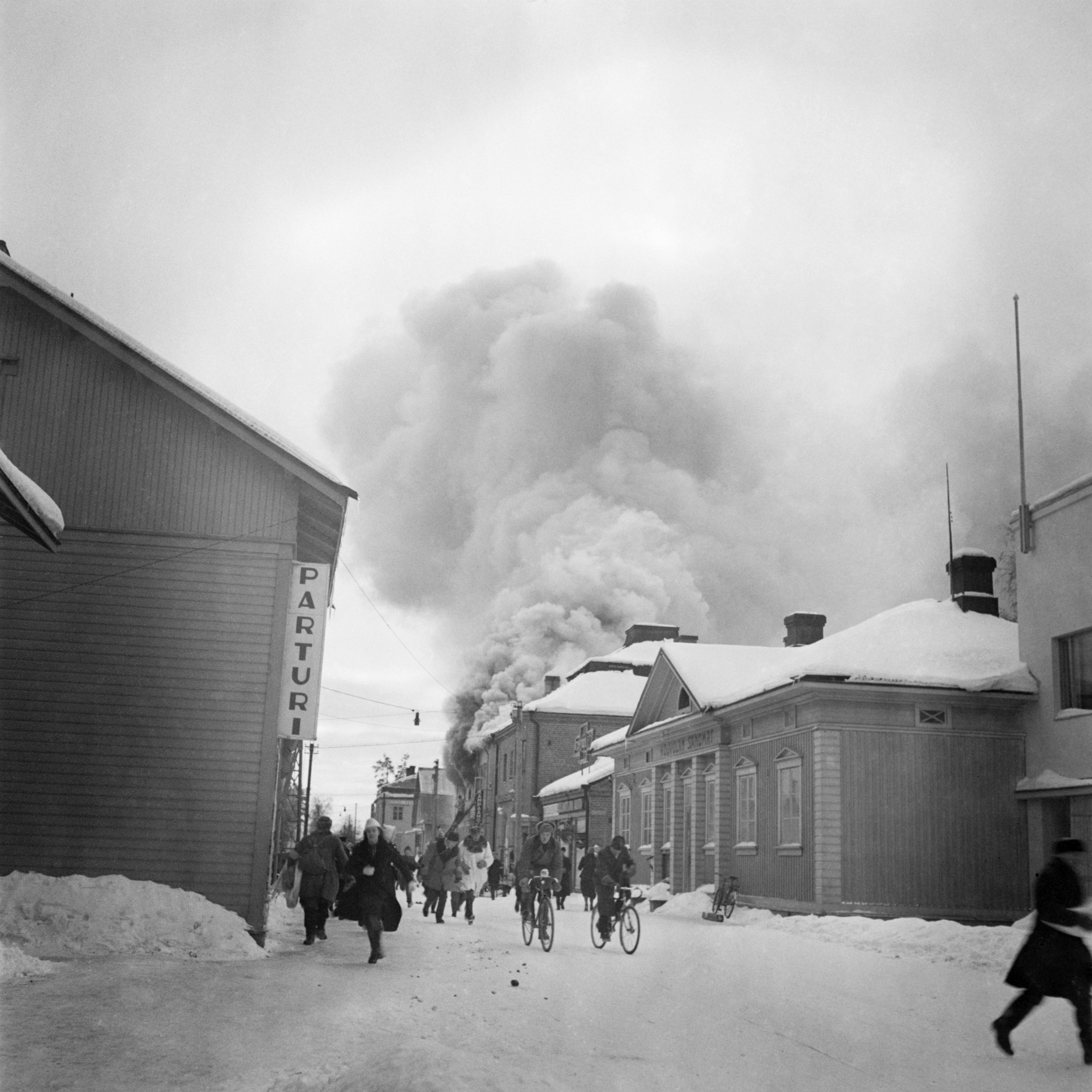 hmiset pakenevat turvaan Neuvostoliiton pommituksilta. Palopommi on sytyttänyt rakennuksen. Oikealla Kouvolan Sanomien toimitus.