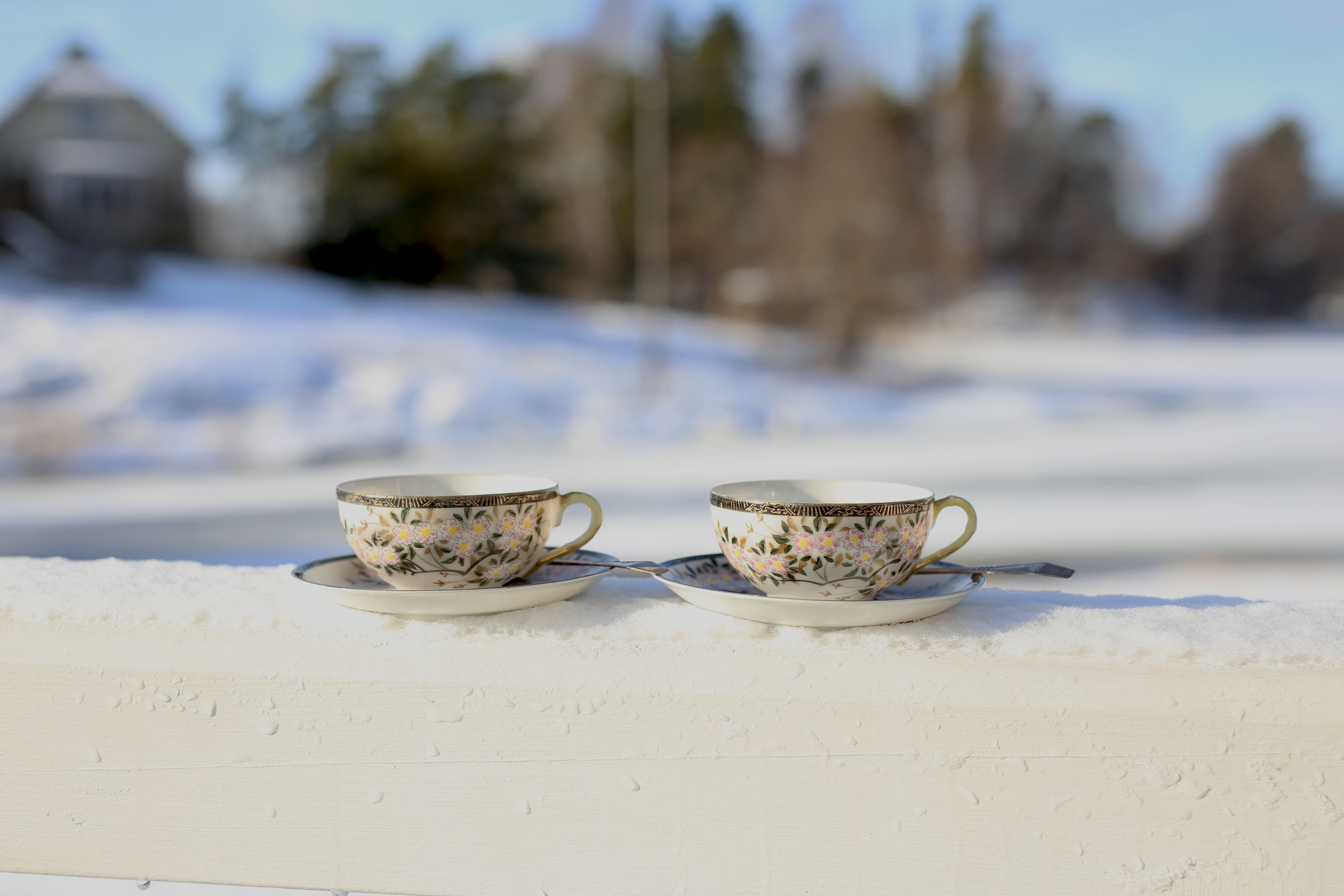 Lumisella kaiteella on kaksi valkoista koristeellista kahvikuppia lautasilla. Tausta on vaalea ja epätarkka. Aurinko paistaa.