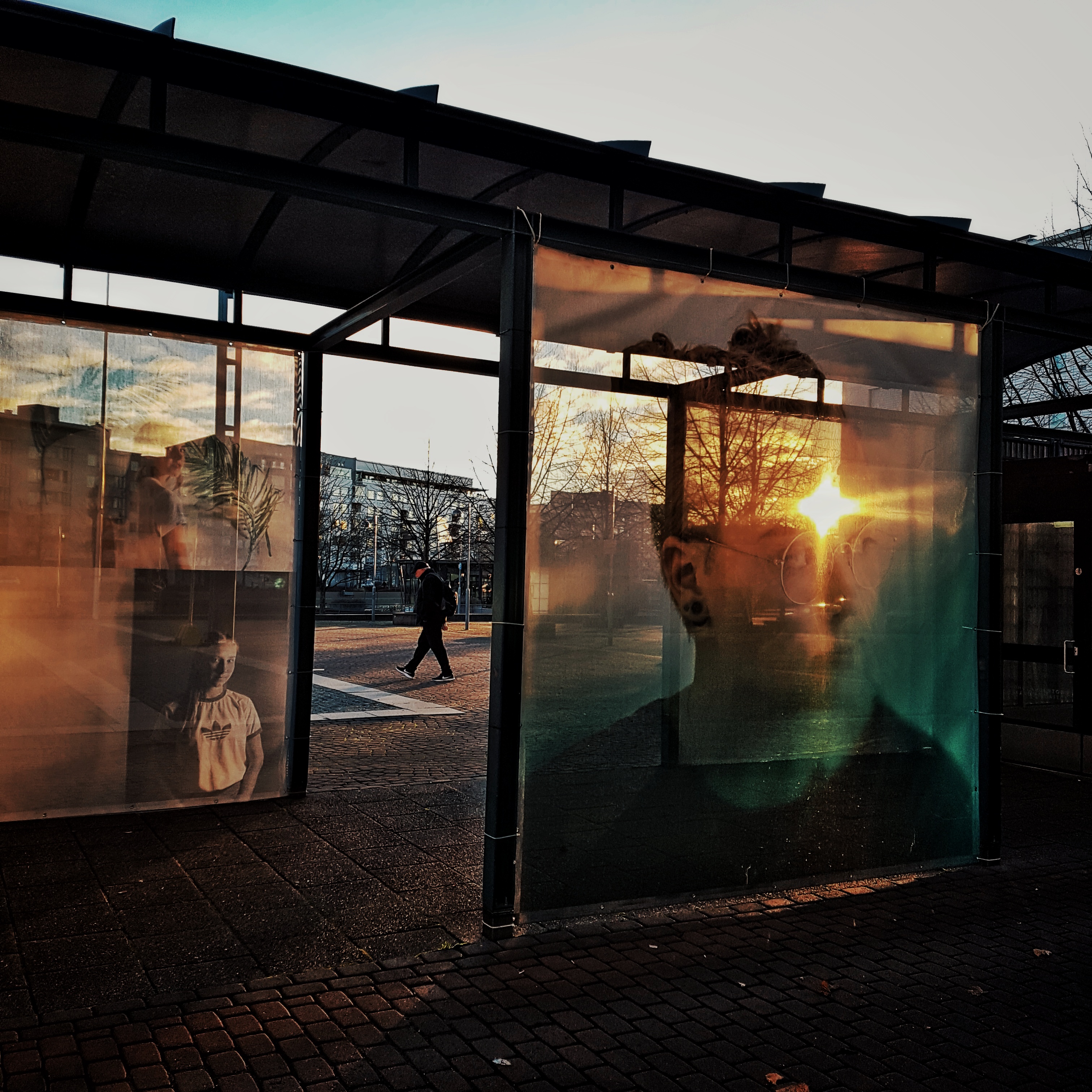 Bussipysäkki tai muu vastaava katos, jonka ilta-aurinko valaisee. Seinissä on haaleita, osin läpinäkyviä kuvia nuorista. 