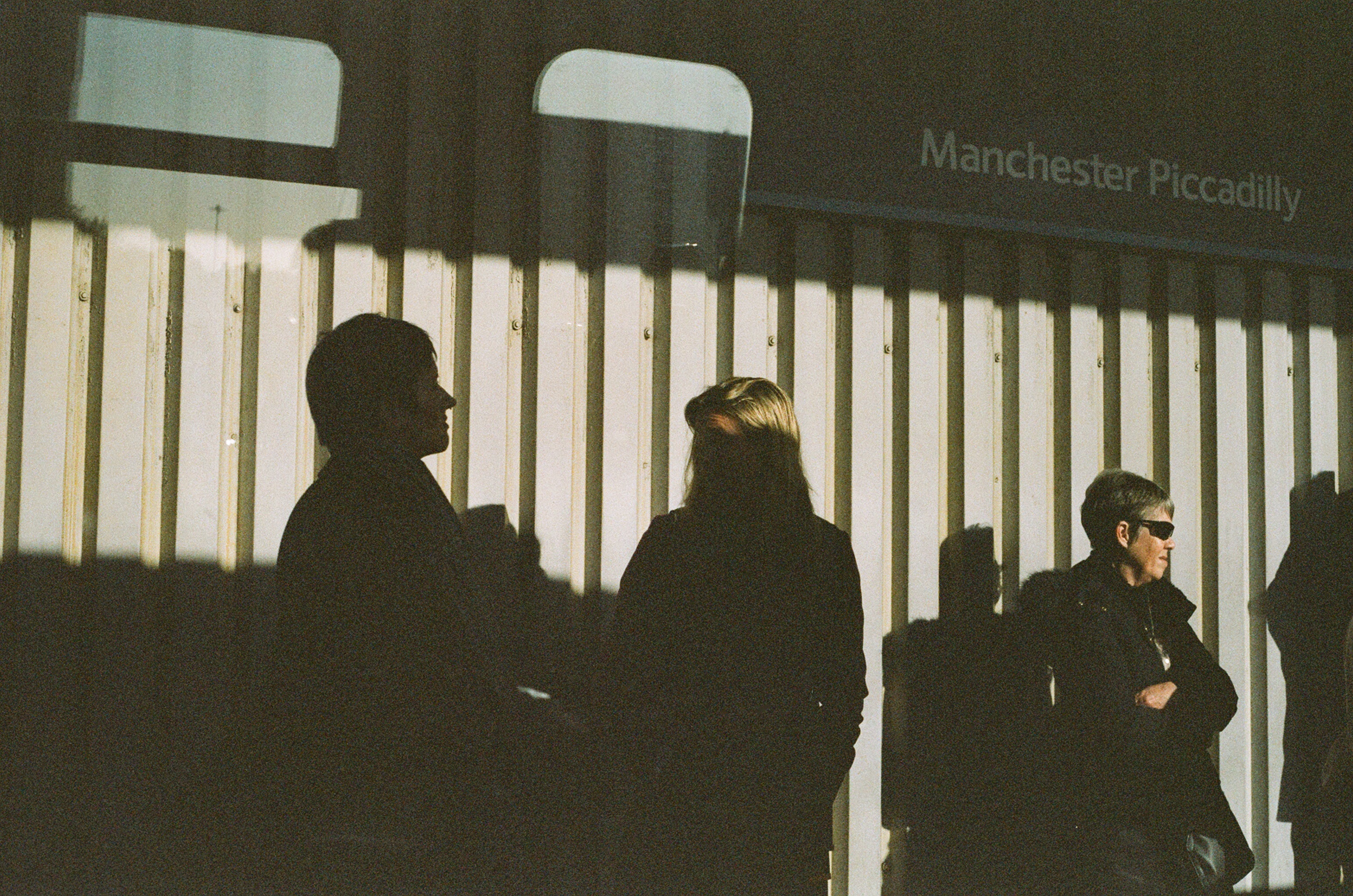 Ihmisiä seisoo seinän edessä. Osa ihmisistä jää varjoon, osa on auringossa. Ylhäällä lukee Manchester Piccadilly.