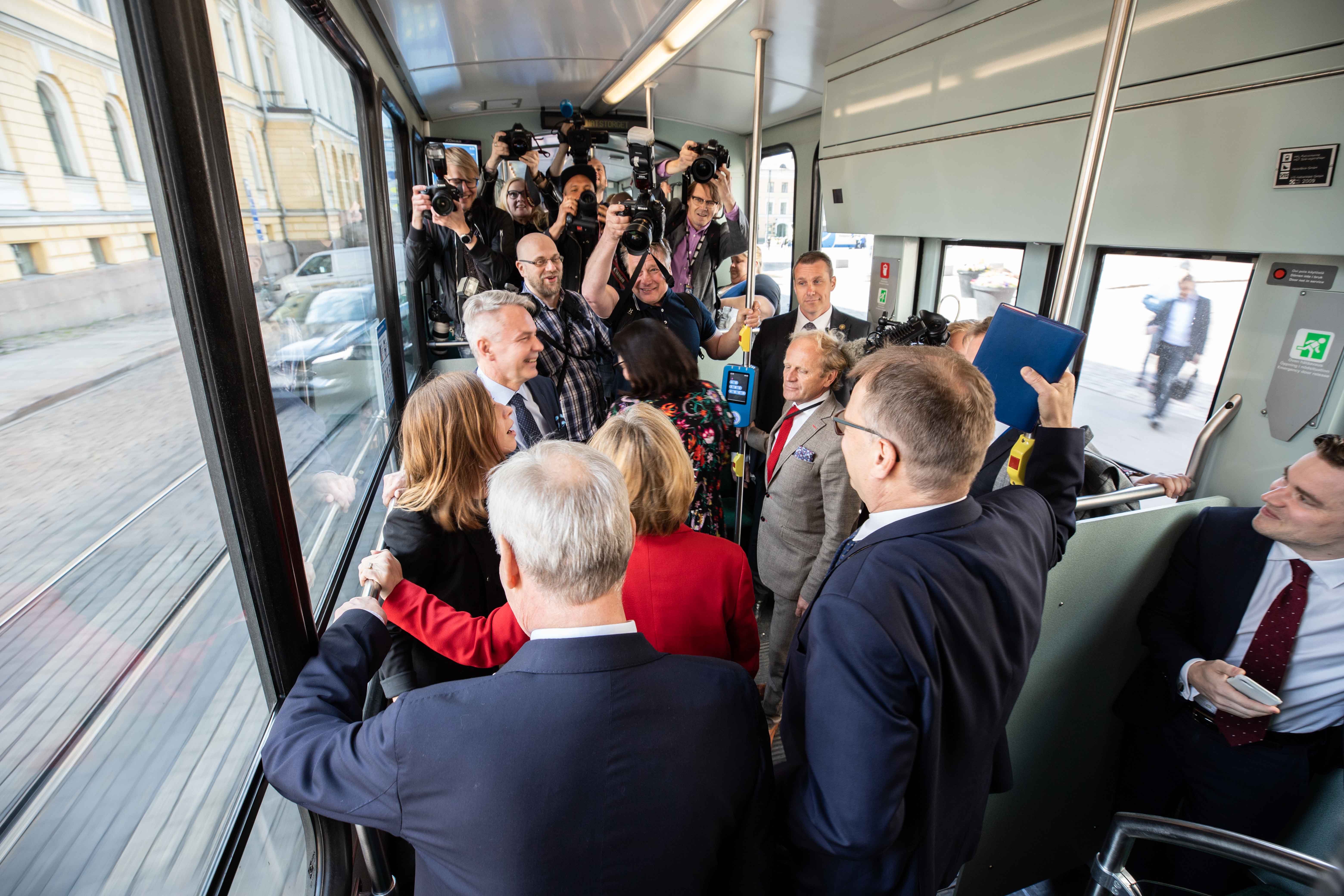 Raitiovaunu täynnä ihmisiä. Etualalla on poliitikkoja puvuissaan, osa selin kameraan. Heidän takanaan on vieri vieressä kuvaajia kamerat kädessään. 