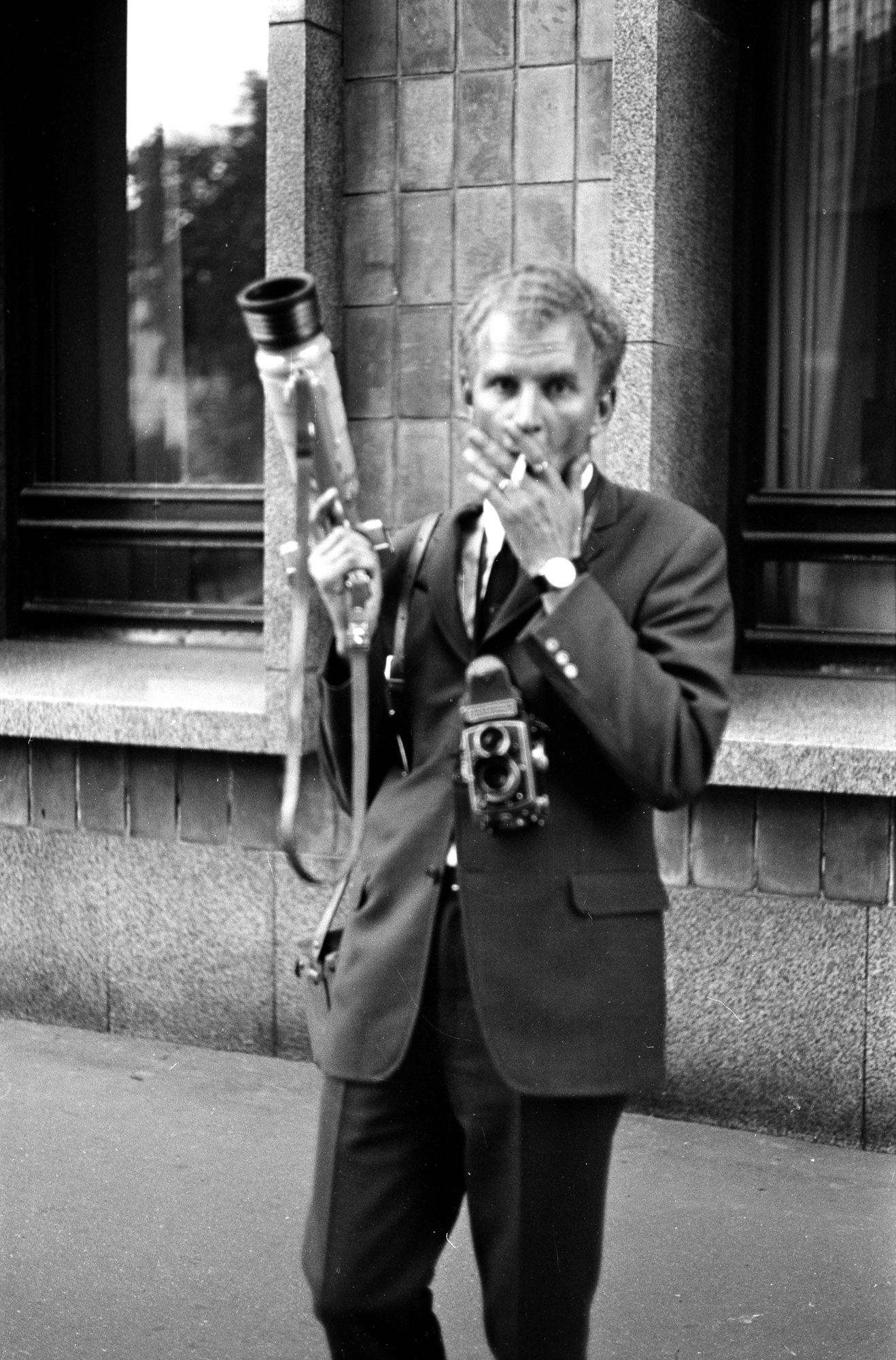 Mustavalkoisessa kuvassa tummaan pukuun pukeutunut mies seisoo jalkakäytävällä rakennuksen edessä, polttaa tupakkaa ja katsoo suoraan kameraan. Hänen kaulastaan roikkuu filmikamera, ja toisessa kädessä hänellä on iso, putkimainen Photosniper-kamera. 
