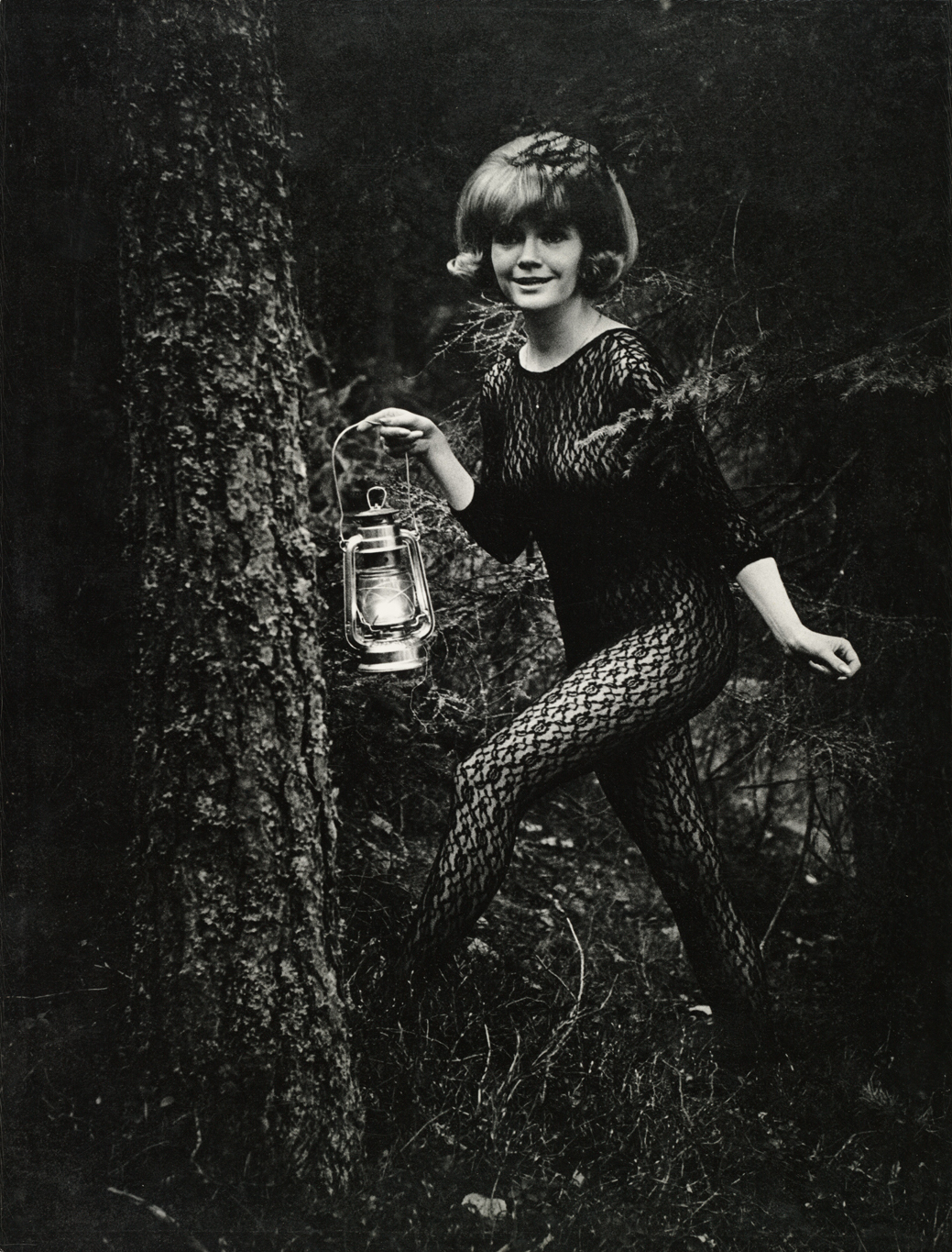 Mustavalkoisessa, tummassa kuvassa nainen poseeraa metsässä kokovartalopitsiasu päällään. Toisessa kädessä hänellä on lyhty.