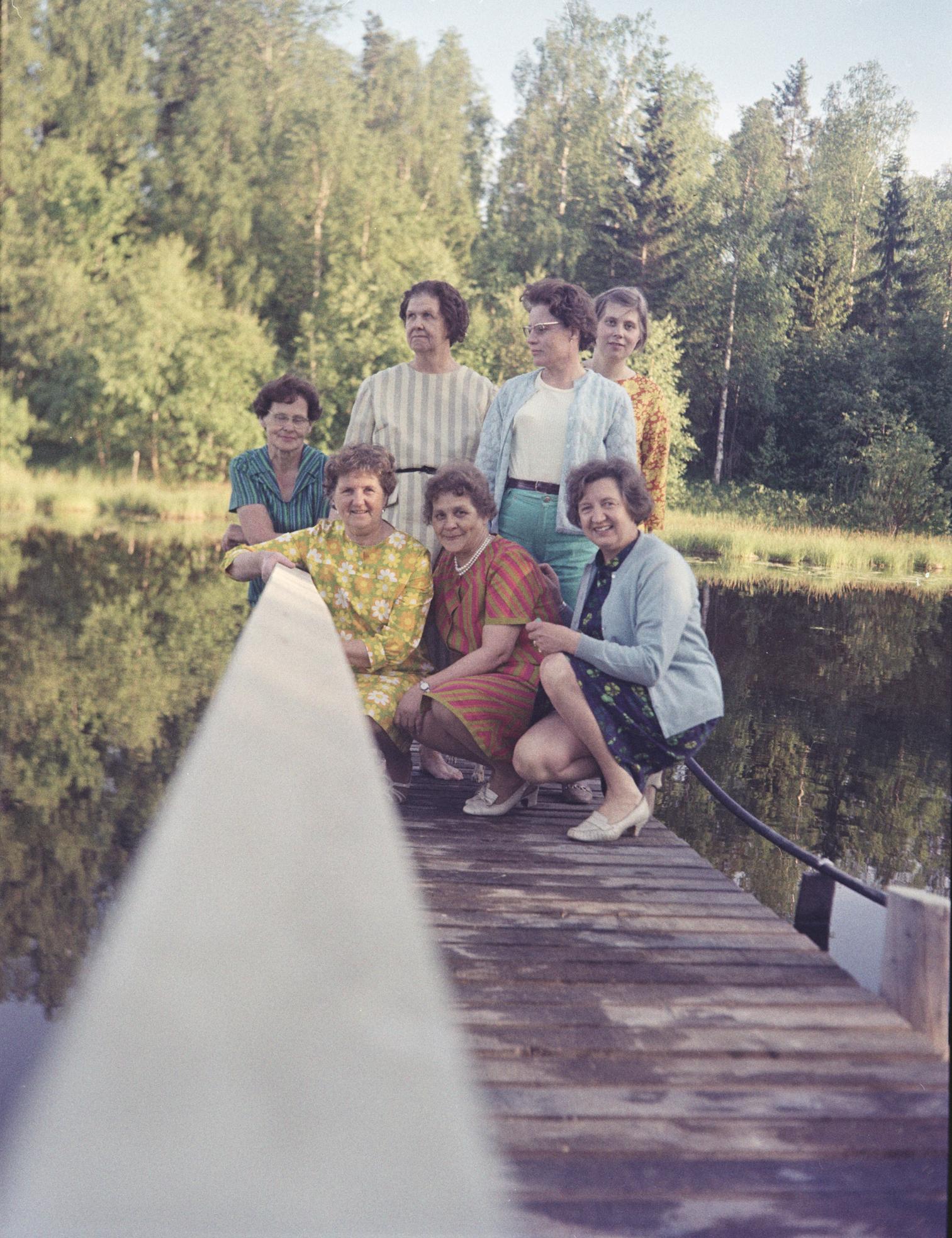 Laiturin päässä on 7 vanhempaa naista. Osa seisoo ja osa on kyykyssä. Laiturin kaide on kuvan keskellä, kuva on luultavasti otettu sitä vasten. Naisten takana näkyy vihreää metsää.