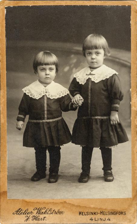 Mustavalkoisessa kuvassa seisoo vierekkäin kaksi pientä lasta. Heillä on samanlaiset vaatteet, musta mekko jossa on pitsikaulus, sekä samanlaiset kampaukset. He pitävät toisiaan kädestä ja katsovat vakavana ainakin lähes suoraan kameraan.