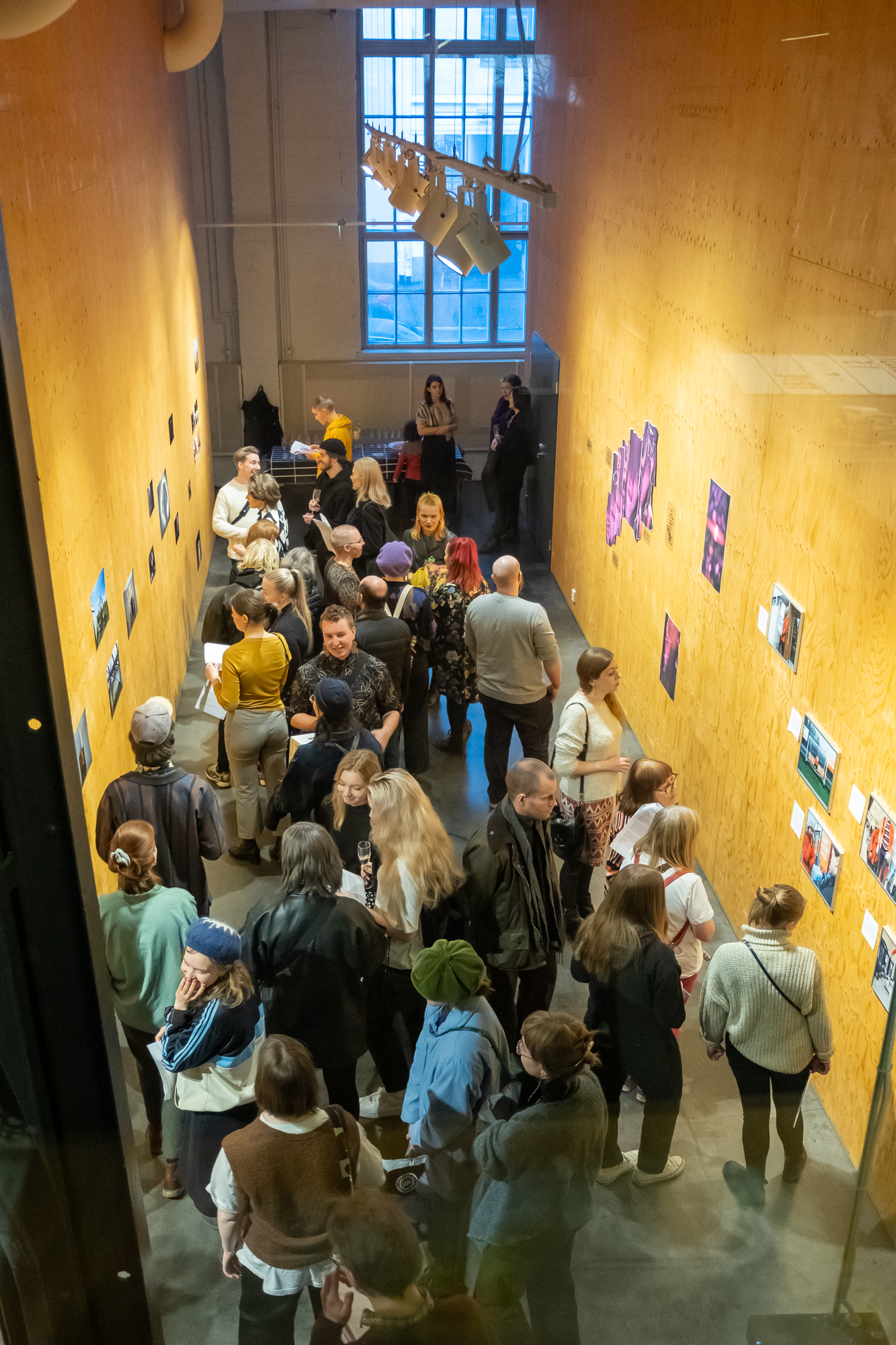 Ylhäältä päin otettu kuva näyttelytilasta, jossa ihmisiä on katsomassa seinille kiinnitettyjä valokuvia. 