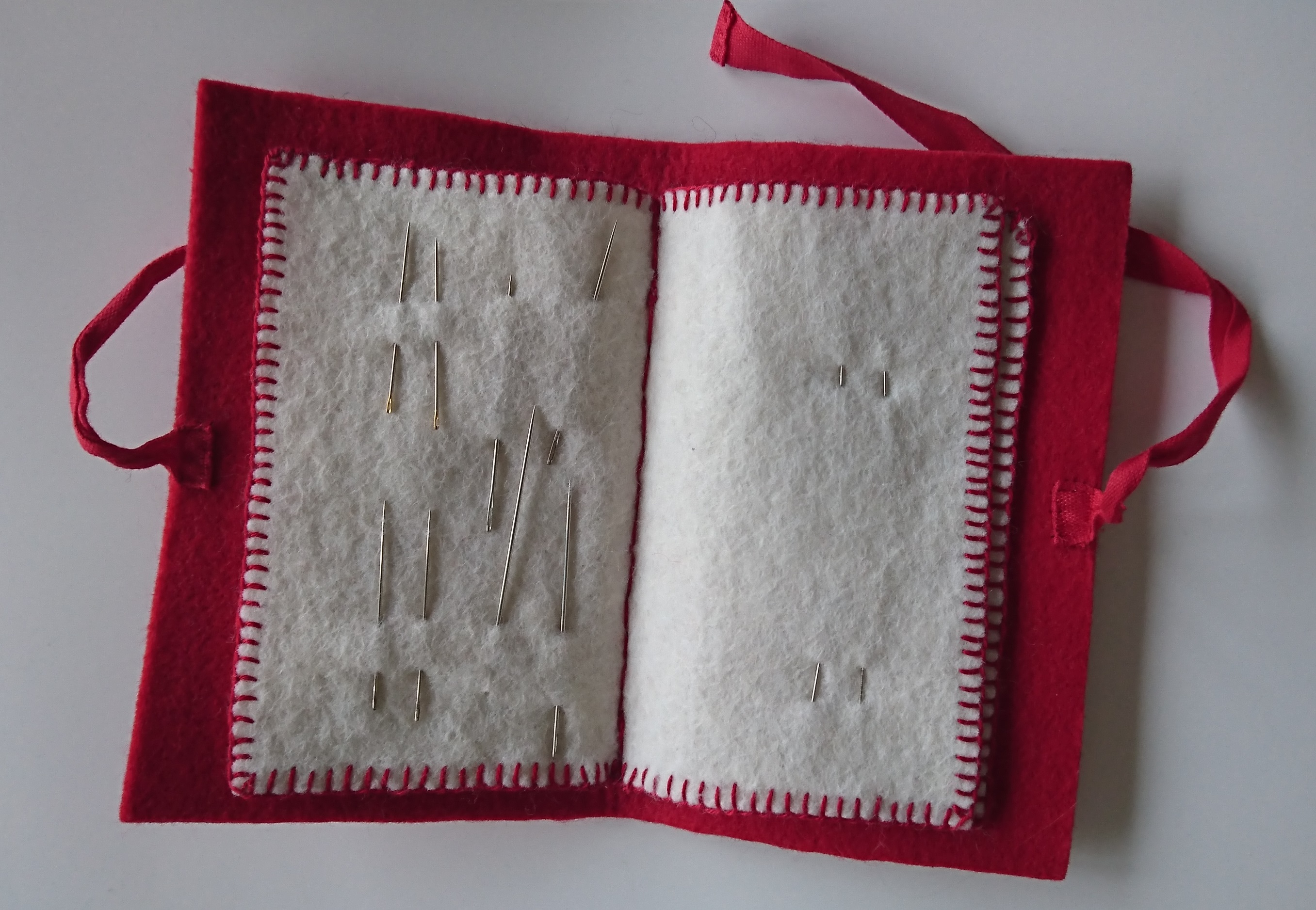 Punaisesta huovasta tehty kirjanen, jossa on "sivut" valkoisesta huovasta. Huopaan on pistetty neuloja. 