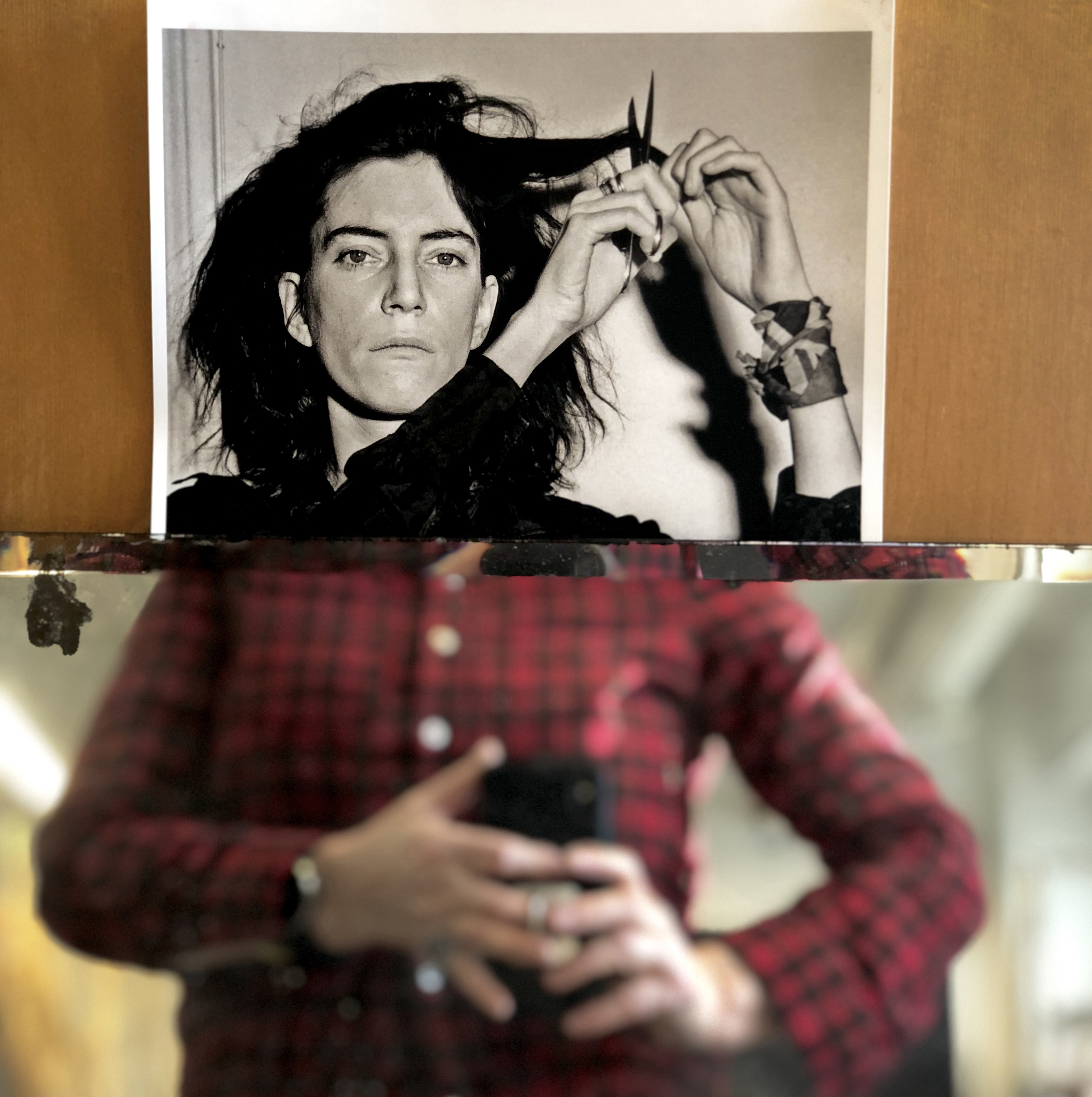 Kaksi kuvaa liitettynä yhteen. Ylemmässä on mustavalkoinen juliste, jossa on nainen juuri leikkaamassa omia hiuksiaan saksilla. Alemmassa on epäterävä naisen keskivartalo, kädet pitävät puhelinta. 