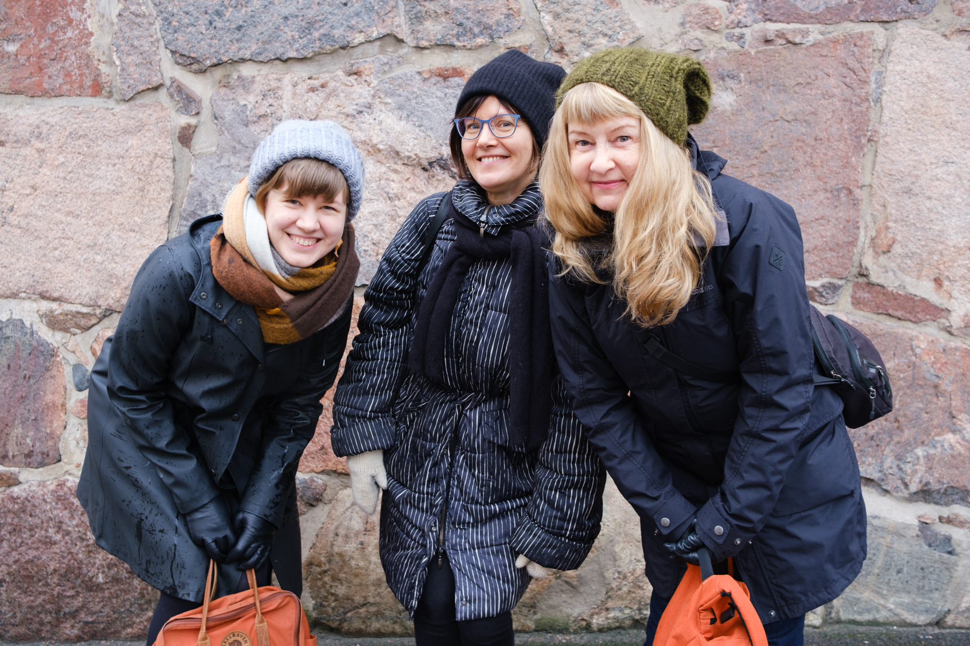 Kolme naista seisoo ulkona kivisen seinän edessä. Kaikki hymyilevät, kaikilla on pitkä takki ja pipo päässä. 