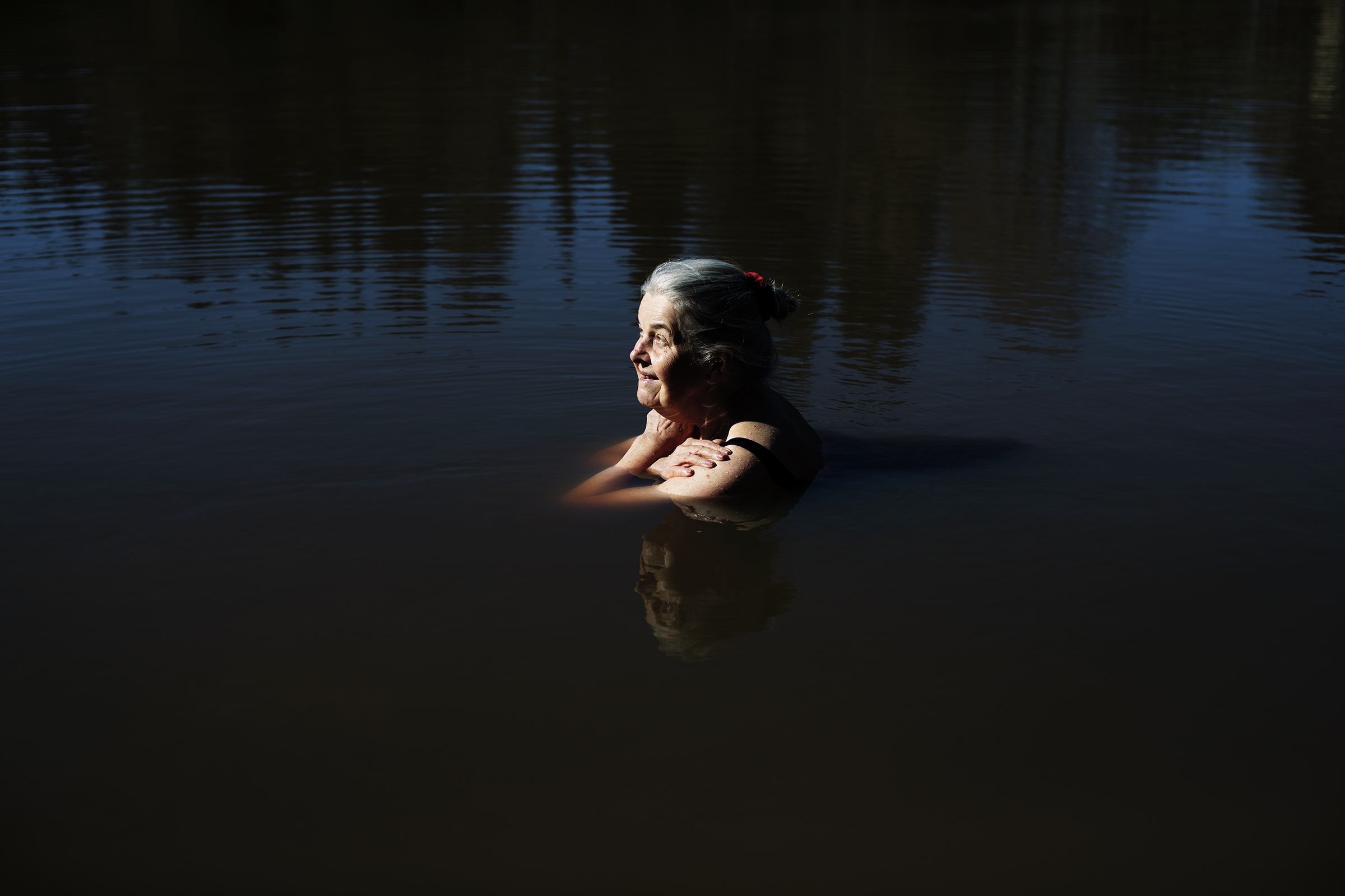 Vanha harmaahiuksinen nainen on vedessä kainaloita myöten. Aurinko valaisee hänet, vedenpinta on tyyni ja tumma.