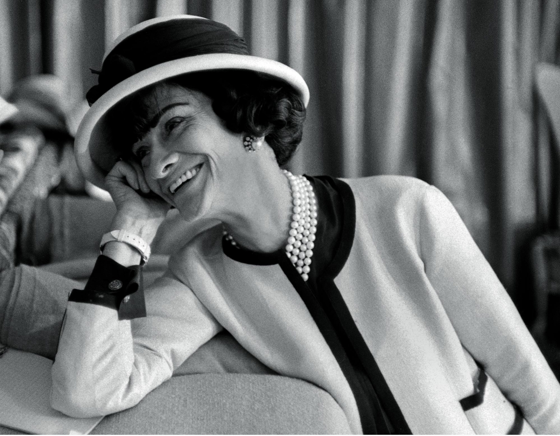 Coco Chanel nojaa käsivarteensa hymyillen. Hän on klassisesti pukeutunut, mukaan lukien hattu, helmet ja rannekello.