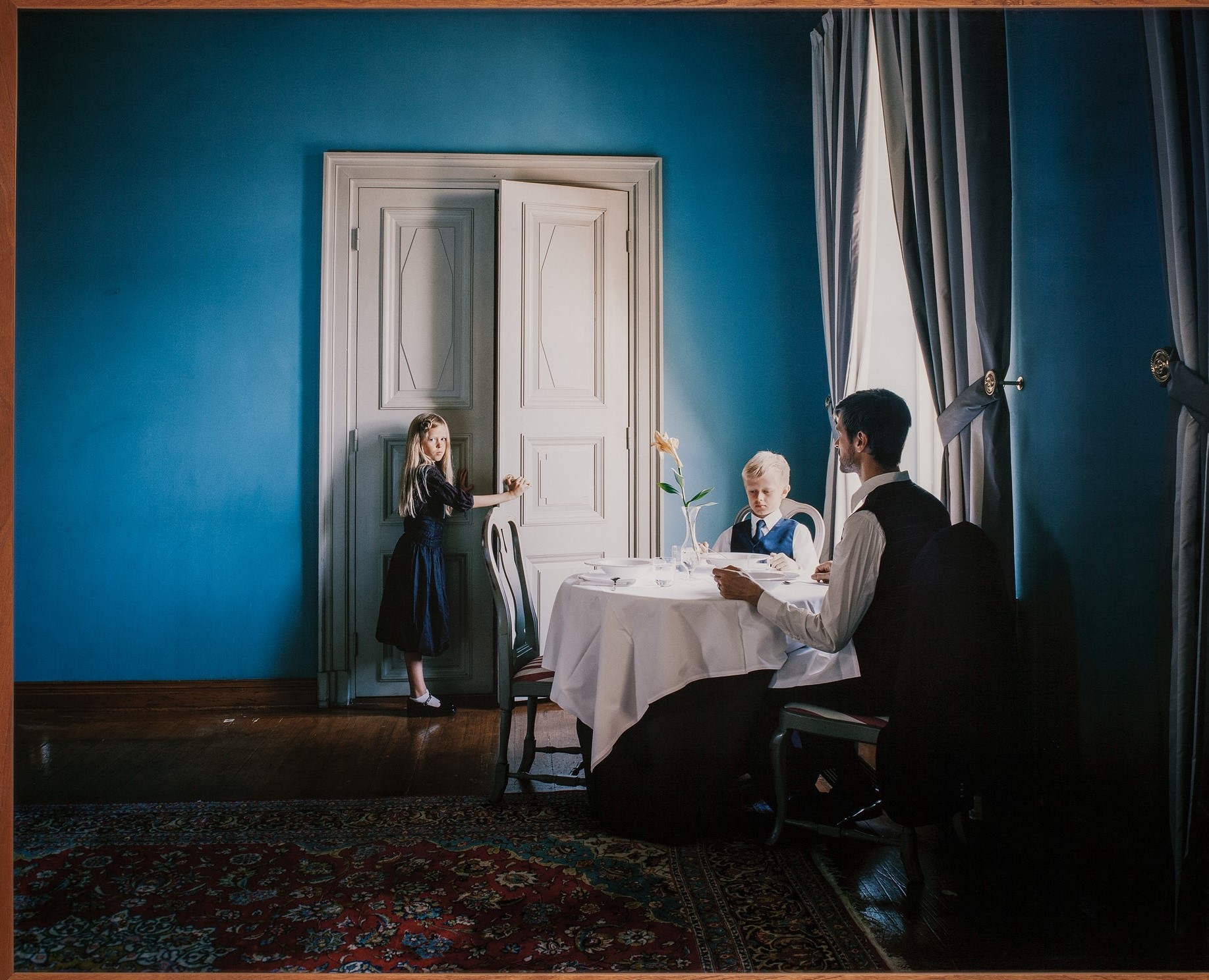 Siniseinäisessä huoneessa mies ja poika istuvat valkoisella pöytäliinalla peitetyn pyöreän pöydän ääressä. Heillä on kauluspaidat ja solmiot. Takimmaisella seinällä on ovi, jota nuori tyttö sinisessä mekossa on avaamassa. Hän katsoo kameraan, mies katsoo tyttöä. 