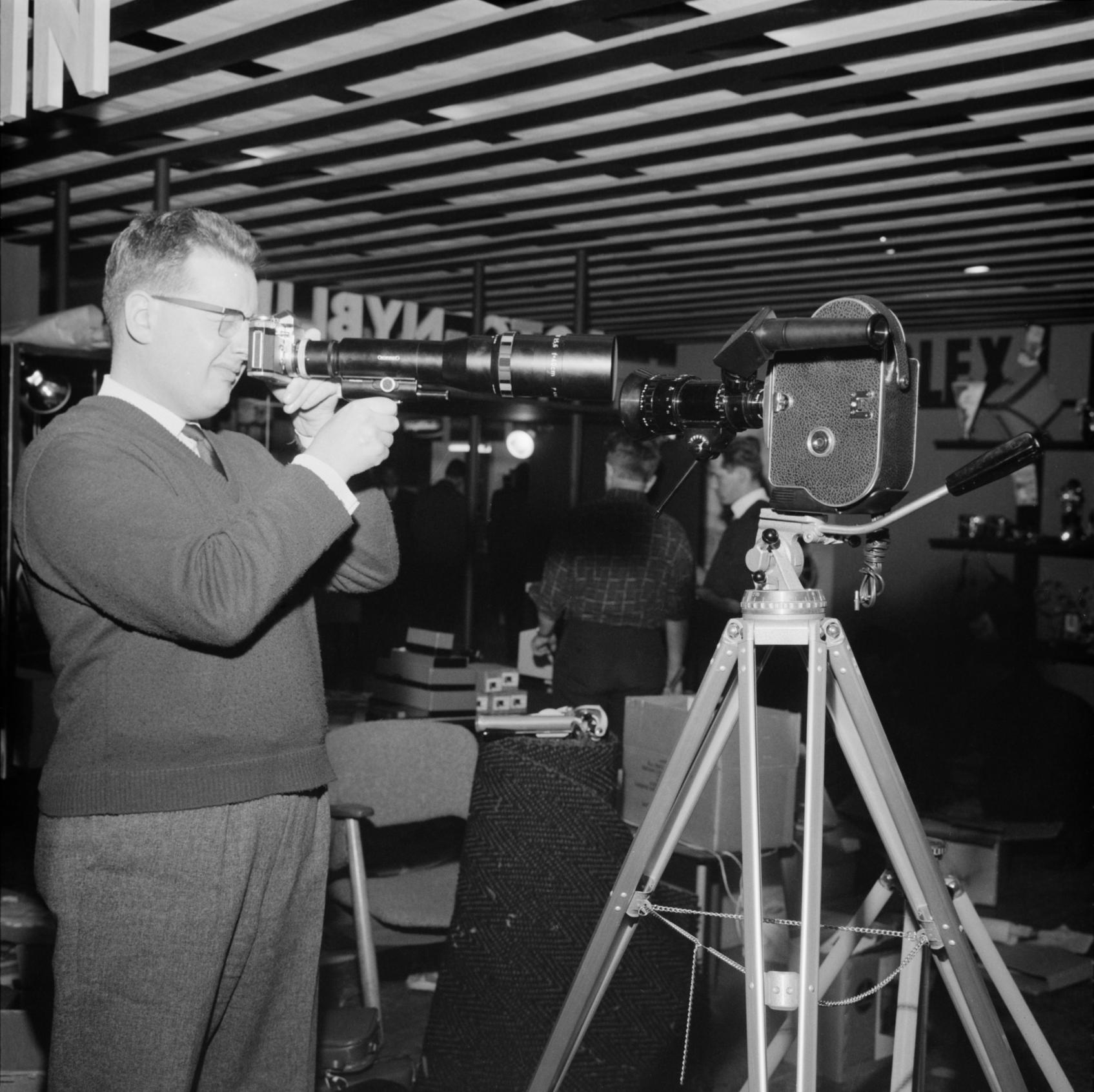 Myyntipäällikkö Reijo Tammi esittelee vuonna 1962 400 mm:n Novoflex-teleobjektiivilla varustettua kameraa. Jalustalla oikealla on 16 mm:n zoom-objektiivilla varustettu elokuvakamera. Uuden Suomen kokoelma. Suomen valokuvataiteen museo.