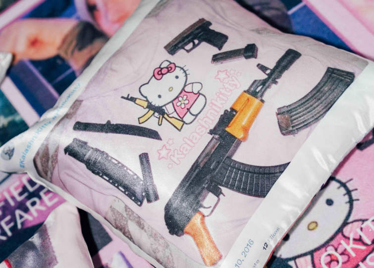 Tyyny, jossa rynnäkkökivääriä pitelevä Hello Kitty -hahmo. Hahmon ympärillä erilaisia aseita. Tyynyn taustalla videopelihahmo ja vaaleanpunaista söpöä estetiikkaa.