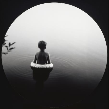 Mustavalkoisessa kuvassa lapsi uimarenkaan kanssa tyynessä vedessä.