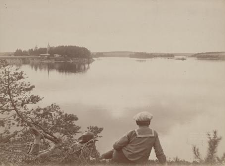 Mustavalkoisen kuvan etualalla on kaksi henkilöä katselemassa järvimaisemaa. 