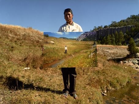 Henkilö seisoo pitäen valokuvaa, jossa on lähes samanlainen maisema kuin missä hän seisoo.