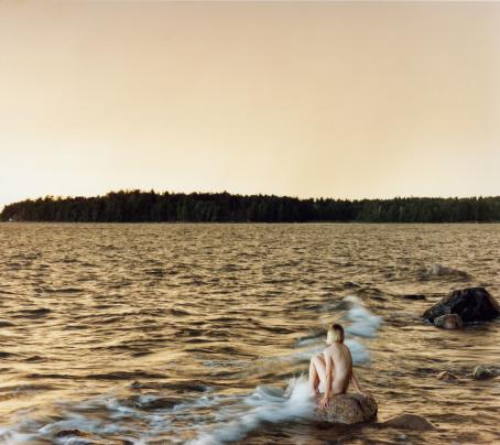 Aallokko, jonka taustalla näkyy metsää. Kivellä istuu alaston ihminen polvet koukussa, aalto lyö hänen jalkoihinsa.