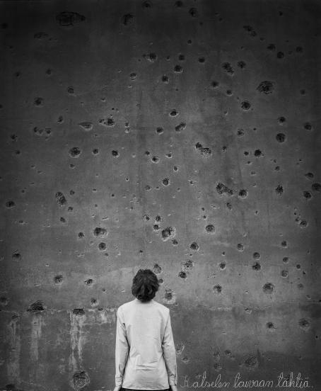 Mustavalkoisessa kuvassa henkilö seisoo katselemassa kiviseinää, jossa on erikokoisia kuoppia ja reikiä. 