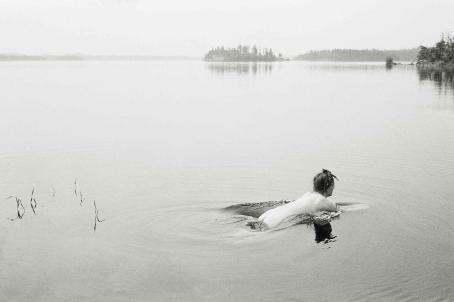 Mustavalkoinen järvimaisema, jossa alaston nainen on juuri kastautumassa veteen.