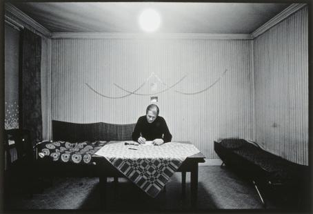 Mustavalkoinen kuva huoneesta. Huoneen keskellä on pöytä, jonka ääressä henkilö istuu kirjoittamassa paperille.