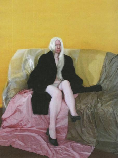 Nainen jolla on pitkät valkoiset hiukset, istuu sohvalla.Hänellä on päällään vain alushousut, tumma turkistakki ja mustat korkokengät. Sohva on peitetty erivärisin kankain ja takana oleva seinä on keltainen.