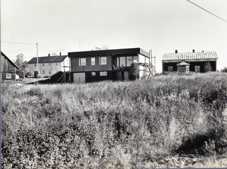 Mustavalkoisessa kuvassa etualalla olevan heinikon takana on rakenteilla suorakulmion muotoinen talo. Molemmilla puolilla sen takana on vanhoja isoja puutaloja.