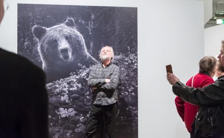 Heikki Willamo poseeraa karhuvalokuvansa kanssa näyttelyssä ja joku ottaa hänestä kuvaa puhelimella.